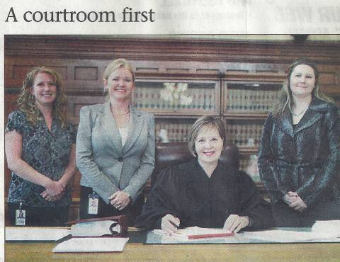 3 women standing, 1 woman seated in courtroom in front of bookcase; left to right are, Michelle Strietbeck, Dawne D. Lindsay, Judge Diane O. Leasure, and Amy McDonald