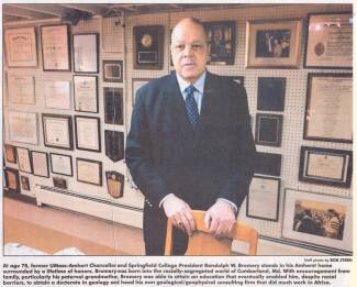 Photo of Randolph Bromery standing with numerous plaques and pictures on wall
