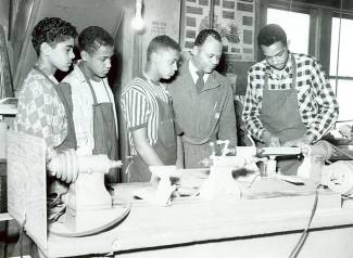 4 Carver High School student with aprons on participate in shop class while teacher oversees woodwork