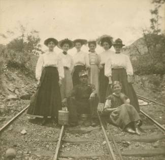 1 miner sitting surrounded by 8 women standing on railroad tracks