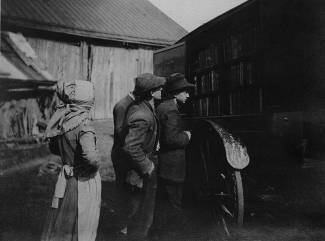 Woman in bonnet with 2 young men standing outside of book wagon, looking over books