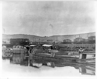Several canal boats docked, train in background; circa 1880