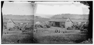 Circa 1862 photo of barn used for hospital; white tents pitched in background, horses and soldiers outside 