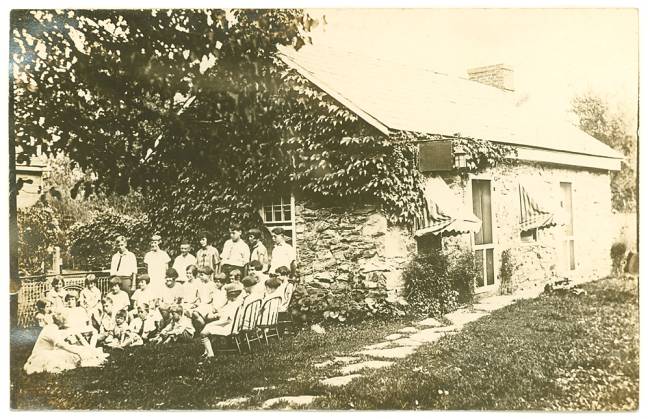 Group of children sitting in countryside outside of a stone home listening to librarian read a story