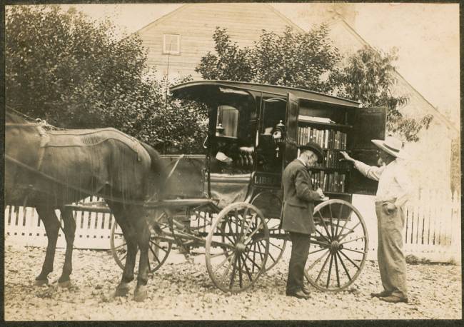 Book wagon pulled by horse with 2 men standing looking through book shelf