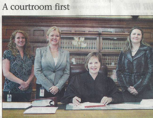 3 women standing, 1 woman seated in courtroom in front of bookcase; left to right are, Michelle Strietbeck, Dawne D. Lindsay, Judge Diane O. Leasure, and Amy McDonald