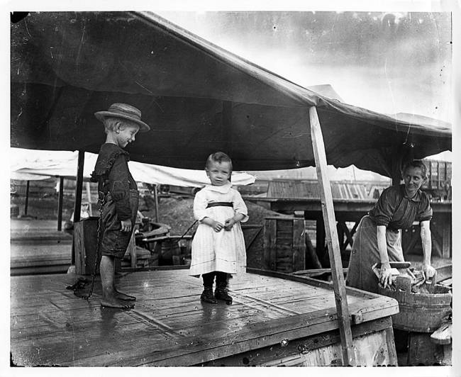 two young children playing on boat wearing safety chains; mother looks on from wash tub and tin