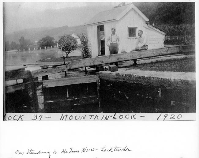 2 men, 1 standing, 1 sitting, by lock gate with shed and trees in background