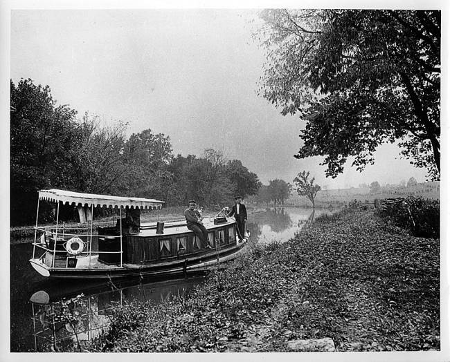 House boat in canal, windows with curtains and cover on front; 2 men at back of boat 