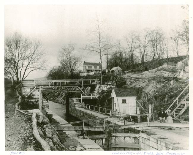 House on hill in distance; lock canal below, lockkeeper and wife stand near the lock gate