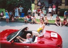 Woman with crown and flowers on red convertible sitting on back waving in parade; man driving