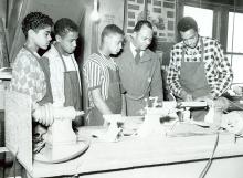 4 Carver High School student with aprons on participate in shop class while teacher oversees woodwork