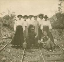 1 miner sitting surrounded by 8 women standing on railroad tracks