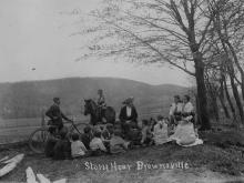 Children sitting on ground on countryside listening to librarian telling story; boy with bike, boy on horse