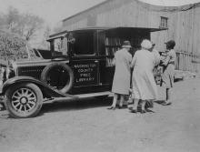 Book wagon parked outside of barn in country; 4 people look through books outside