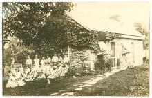 Group of children sitting in countryside outside of a stone home listening to librarian read a story