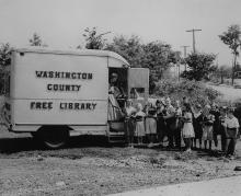 Bookmobile in 1950s parked on side of road; children standing outside holding books