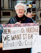 Photo of Mary Miltenberger holding a sign "The Monument is Our Heritage We Want it Back in Front of Our Courthouse"