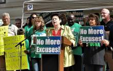 Women gather at podium during correctional guard rally, 2012 holding signs "No More Excuses"