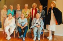 Photo of 9 women; 6 standing 3 sitting with mannequin dressed in nursing attire 