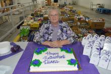 Photo of Diana Loar with a cake "Happy Retirement Diana" with boxes of food pantry items in background