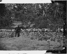 Picture of solider standing by stone wall with 12 stones in ground for grave markers