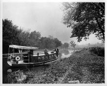 House boat in canal, windows with curtains and cover on front; 2 men at back of boat 