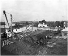 Coal is being lifted by a steam loader from the boat into waiting horse-drawn wagons; train tracks at bottom, canal at left