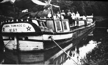 crowd of people stand on the deck of the Canal Towage boat 67, tied up along the canal