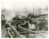 House on hill in distance; lock canal below, lockkeeper and wife stand near the lock gate